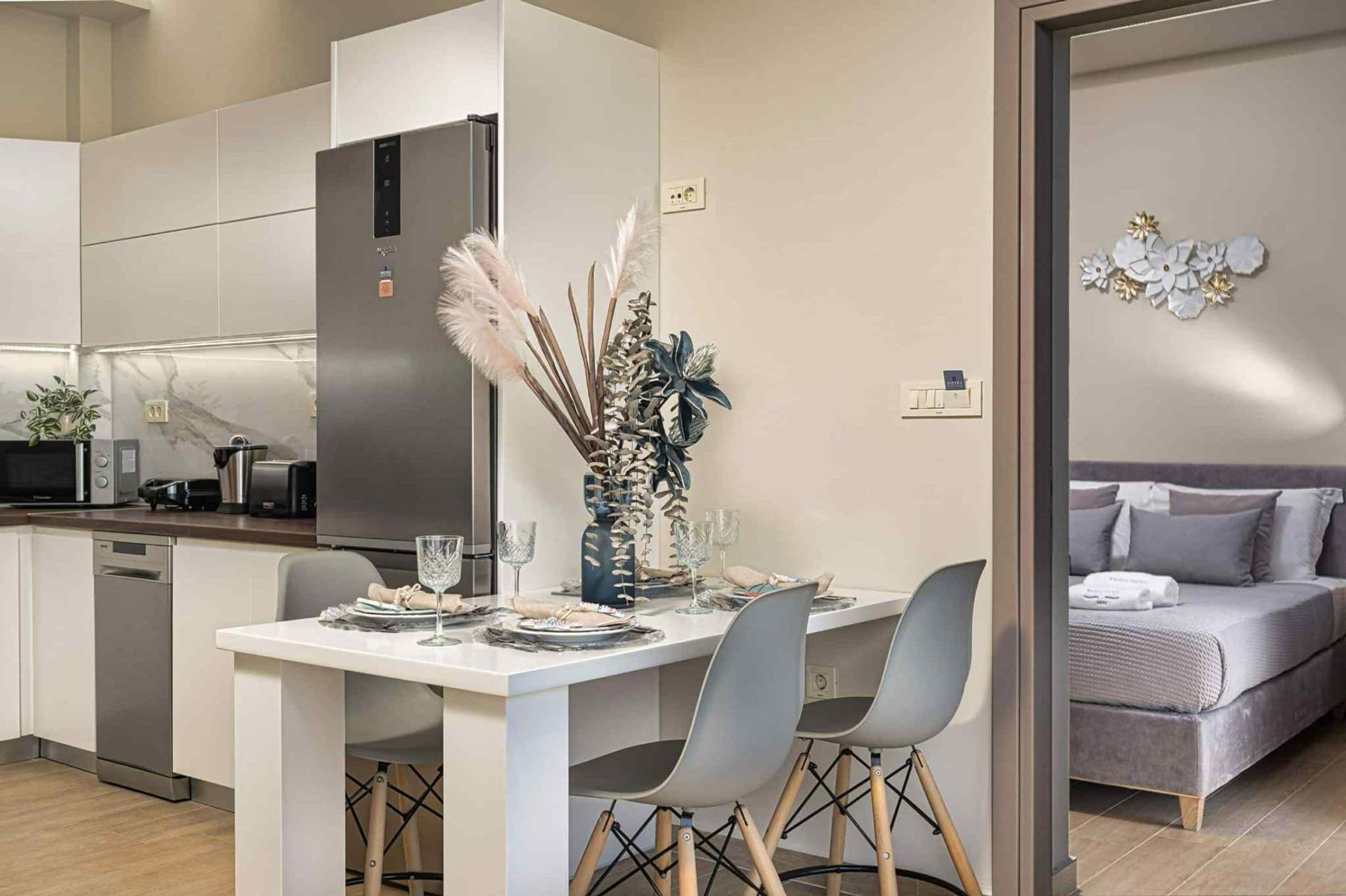 Una cocina moderna con refrigerador integrado, una mesa de comedor preparada para dos personas y un sofá gris en la zona de estar contigua.