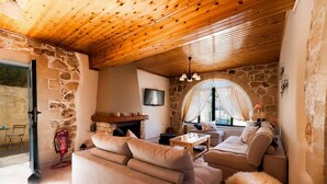 A cozy living room with a stone wall, wooden ceiling, and a fireplace. There is a sofa, a coffee table, and a television mounted on the wall. - Villa Kirke in Chania (Apokoronas)