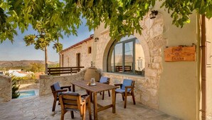 A stone patio with a wooden table and chairs, a stone wall, and a view of the landscape. - Villa Kirke in Chania (Apokoronas)