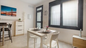 Un coin repas avec une table dressée pour deux personnes, une chaise, un petit meuble et une fenêtre avec des stores.