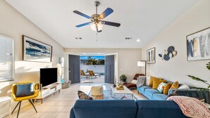 A spacious living room with a blue sofa, a television, and a ceiling fan. - Desert Fun 4BR Retreat | Pool, Jacuzzi & Game Room (Bermuda Dunes)