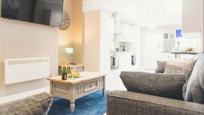 Ein modernes Wohnzimmer mit einem Flachbildfernseher, einem Couchtisch aus Holz mit Flasche und Glas, einem grauen Sofa und einem blauen Teppich.