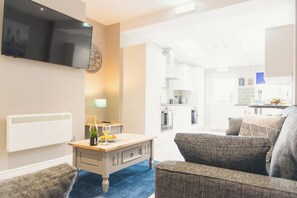 A living room with a flat-screen TV, a clock, a bottle of champagne, and a table.