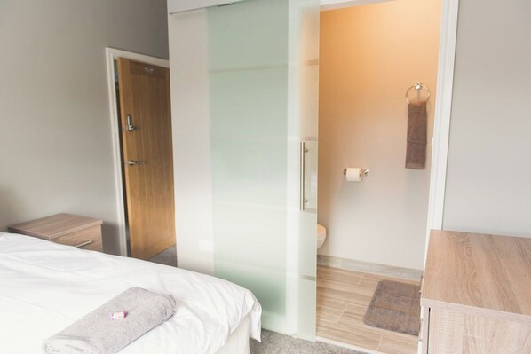 A hotel room with a bed, a wooden nightstand, a glass partition, a bathroom with a toilet, and a towel rack.