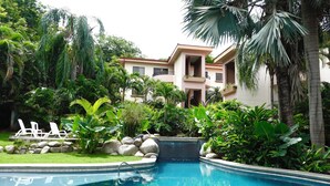 Una piscina con agua azul cristalina, rodeada de exuberante follaje y palmeras. Hay sillas de descanso blancas y un edificio al fondo.