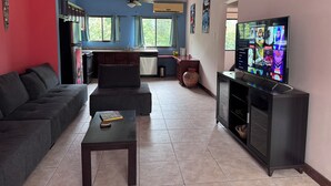Una sala de estar con un sofá, una mesa de centro y un televisor. Hay una zona de cocina con un refrigerador, estufa y alacenas.