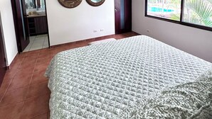 A bedroom with a bed, ceiling fan, two wall mirrors, and a window with a view of greenery. - Experience a perfect vacation in our newly renovated 3-bedroom townhome in Playas Del Coco (Playas Del Coco)