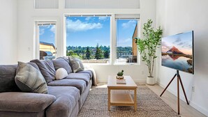 A living room with a gray sofa, a small wooden table, and a TV on a tripod. There are three large windows with a view of a cityscape and a plant on the right.