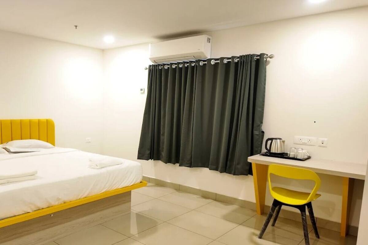 A hotel room with a bed, a desk, a chair, an air conditioning unit, and a curtain.