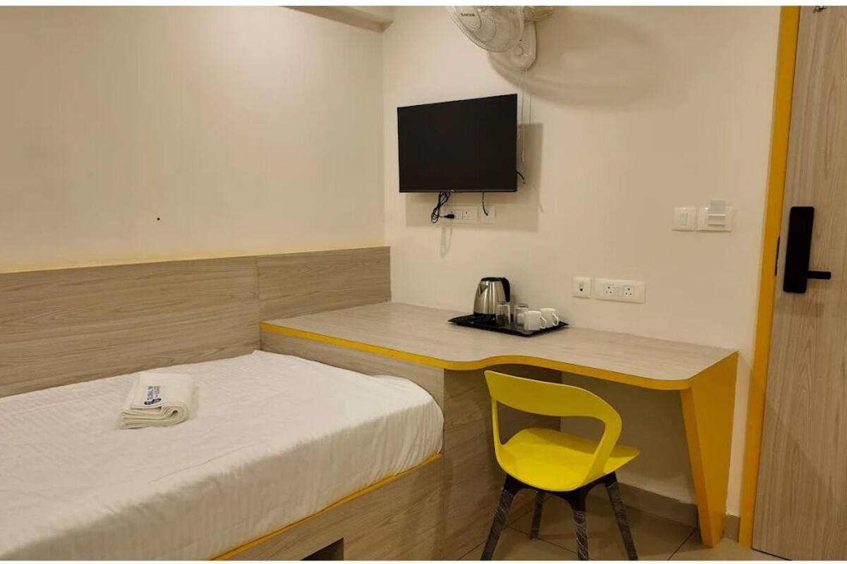 A single bed with a white mattress and a folded towel, a small desk with a chair, a flat-screen TV, and a power outlet.