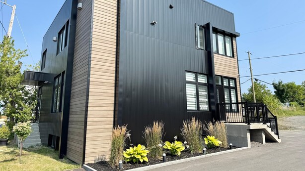 Een modern gebouw met twee verdiepingen, een combinatie van donkere en lichte bekleding, grote ramen en een klein buitenterrein met gele planten.