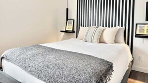 A modern bedroom with a bed, a headboard with vertical lines, and two bedside tables with framed pictures. - Lévis Center | New Studio with Queen Bed + A/C (Levis)