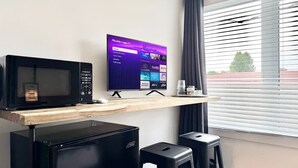 A modern room with a flat-screen TV on a wooden shelf, a microwave, a mini-fridge, and two black stools. - Lévis Center | New Studio with Queen Bed + A/C (Levis)