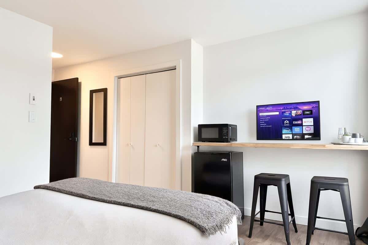 Quarto de hotel moderno com uma cama, uma escrivaninha com monitor de computador, micro-ondas e dois banquetes.
