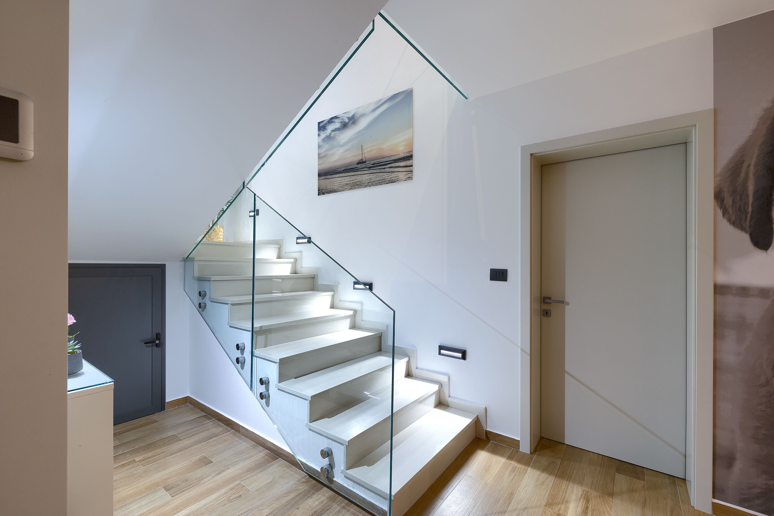 A modern staircase with glass railings, a painting of a sunset, and a wooden floor.