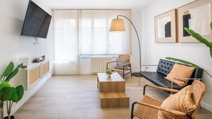Un salon moderne avec un téléviseur à écran plat, un canapé, une table basse et un fauteuil. Il y a une grande fenêtre avec des rideaux légers, une lampe sur pied et un radiateur.