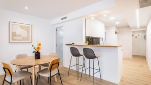 Une cuisine moderne avec des armoires blanches, un plan de travail en bois et un four encastré. On y trouve un coin repas avec une table et des chaises en bois, ainsi qu’un petit îlot avec des tabourets de bar.