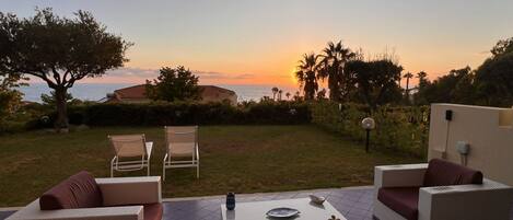 A patio with outdoor furniture, a dining table, and a view of the sunset over the sea.