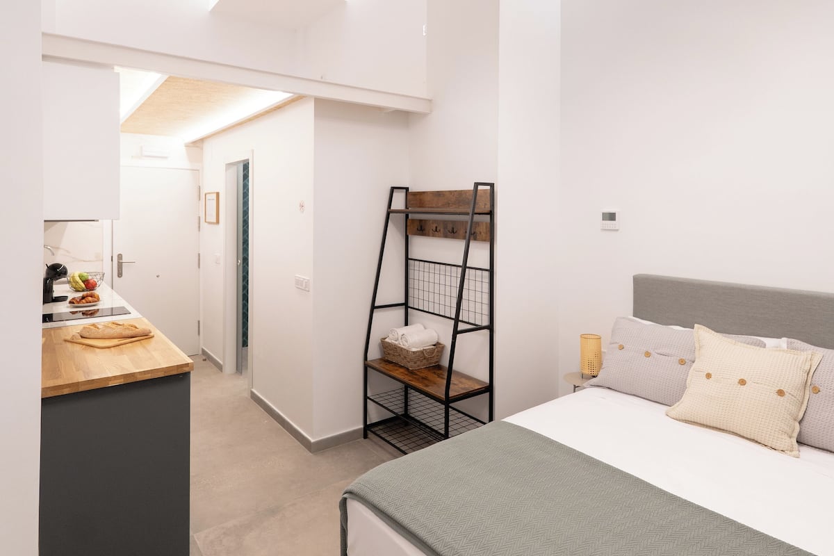 Ein modernes Schlafzimmer mit einem großen Bett, einem Nachttisch und einem Schreibtisch mit Stuhl. An der Wand lehnt eine Leiter, und eine Tür führt in einen anderen Raum.