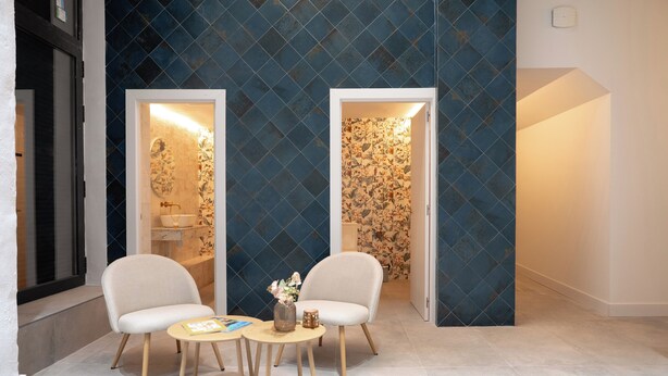 Un baño moderno con un muro azul estampado, dos sillas blancas, una mesita redonda y un florero con flores.