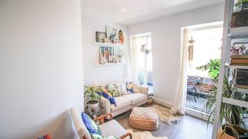 A cozy living room with a sofa, armchairs, and a coffee table. There are shelves with books and decorative items. A large window provides natural light.