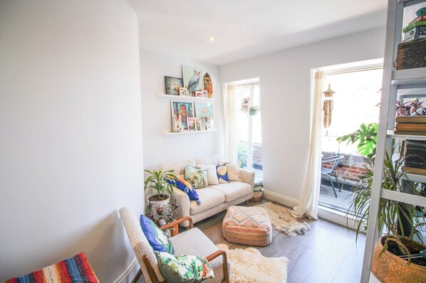 A cozy living room with a sofa, armchairs, and a coffee table. There are shelves with books and decorative items. A large window provides natural light.