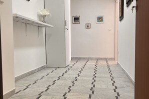 Un pasillo con suelo de baldosas, una puerta blanca y un estante con un florero.