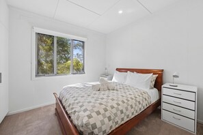 Un dormitorio con cama, mesita de noche, lámpara y ventana con vistas a árboles.