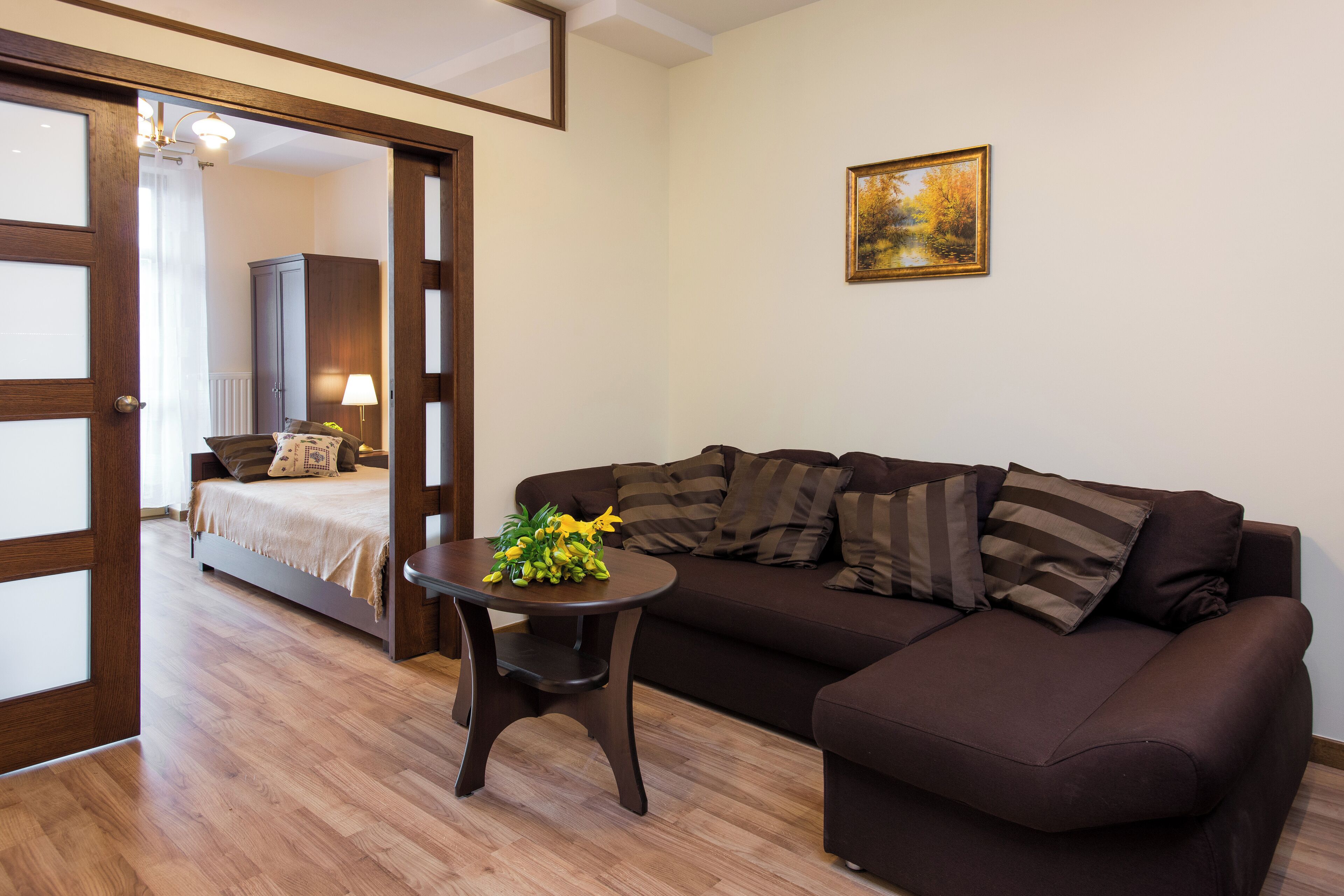 Een kamer met een bruine bank, een klein rond tafeltje met een bloemstuk, een bed met een wit hoofdeinde, en een schilderij aan de muur.
