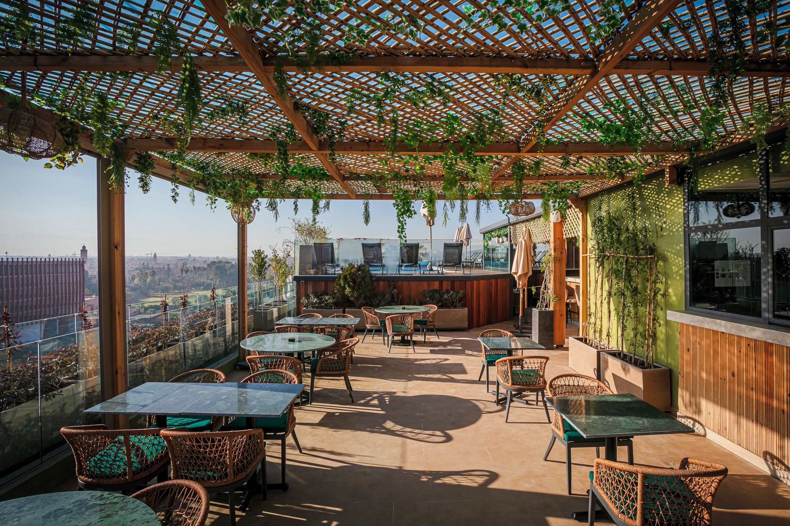 An outdoor patio with tables and chairs, a wooden pergola with greenery, and a view of the cityscape.