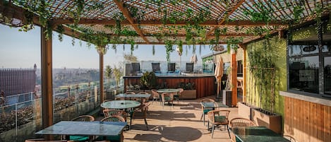 An outdoor patio with tables and chairs, a wooden pergola with greenery, and a view of the cityscape.