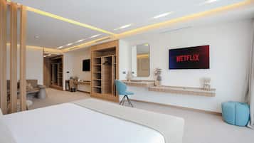 A modern hotel room with a large bed, a seating area, a wardrobe, a mirror, a TV displaying 'NETFLIX', and a small table with decorative items.