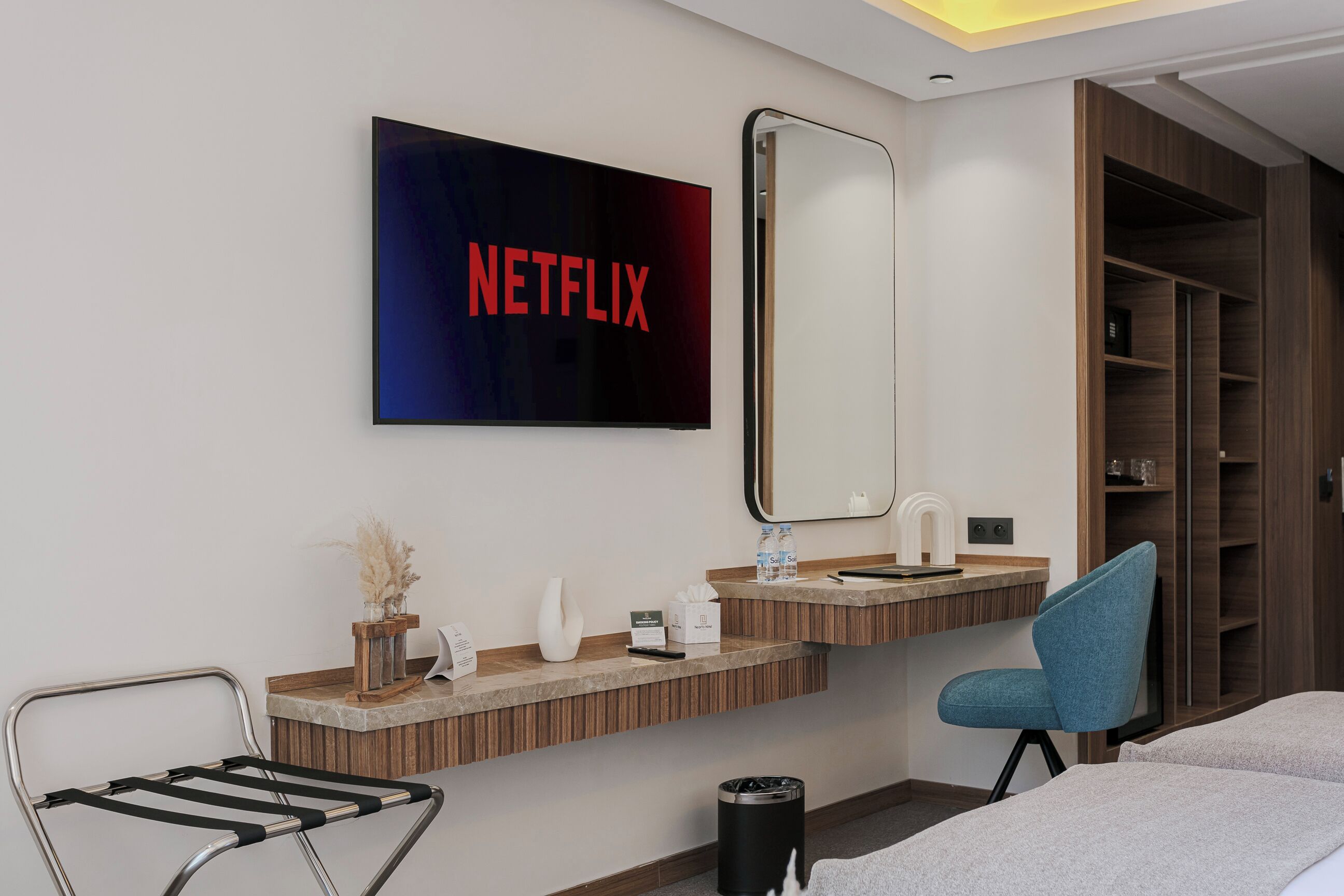 A hotel room with a large mirror, a sink, a chair, a bed, and a wall-mounted television displaying the Netflix logo.