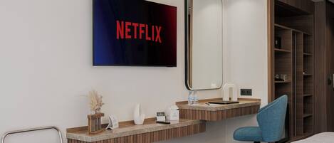 A hotel room with a large mirror, a sink, a chair, a bed, and a wall-mounted television displaying the Netflix logo.