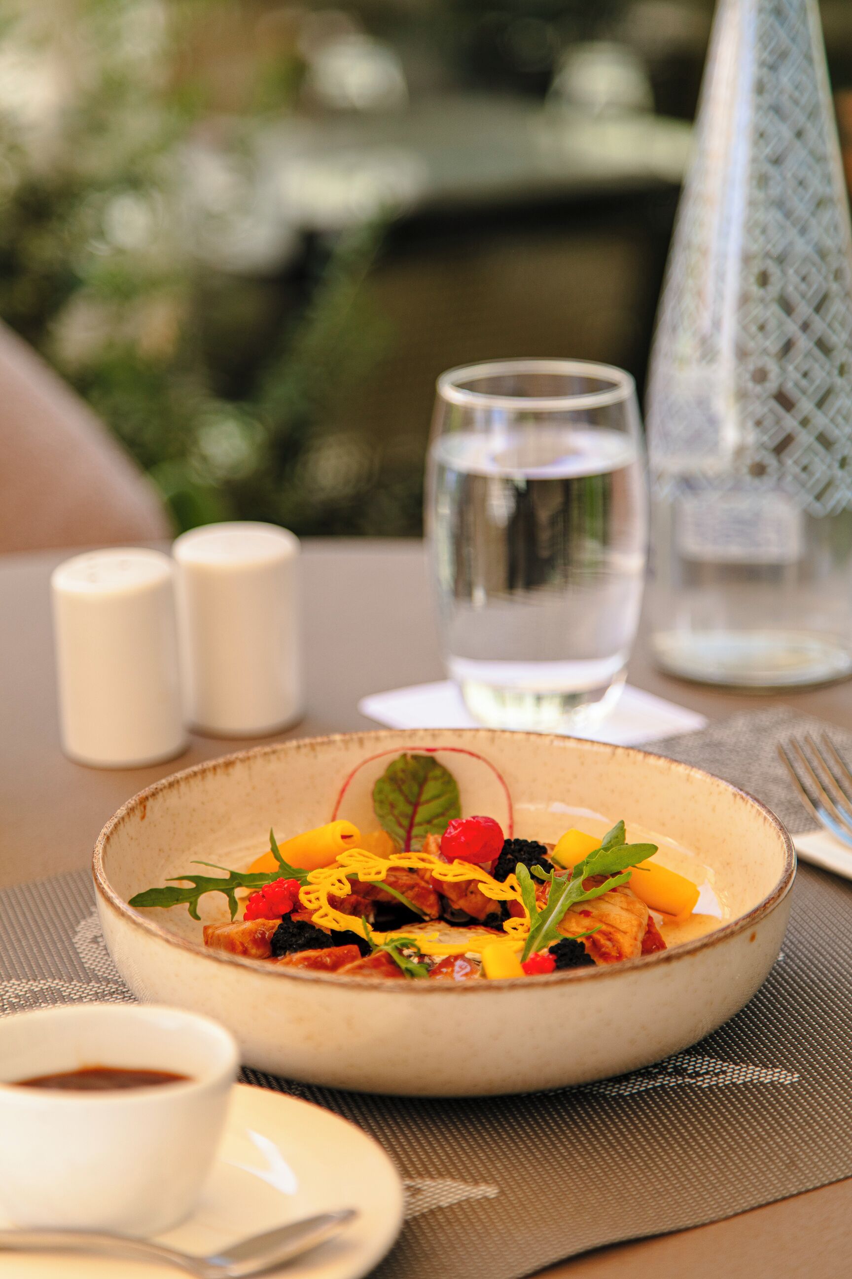 Una mesa de comedor con un tazón de ensalada, un vaso de agua, saleros y pimenteros, una taza de café y un tenedor sobre un mantel individual.