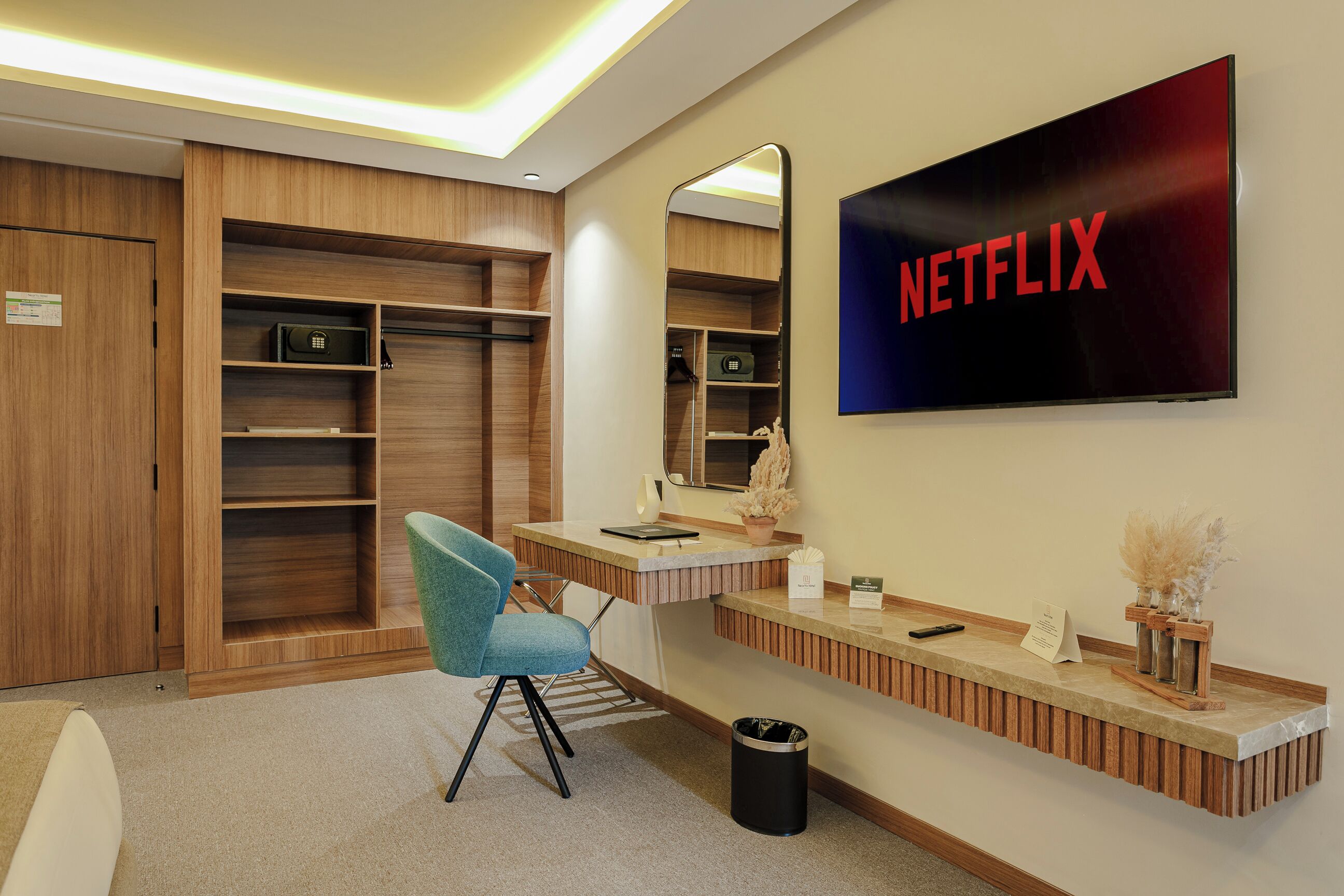Habitación de hotel moderna con tocador de madera, una silla azul, un espejo, un cartel de Netflix y un cubo de basura pequeño.