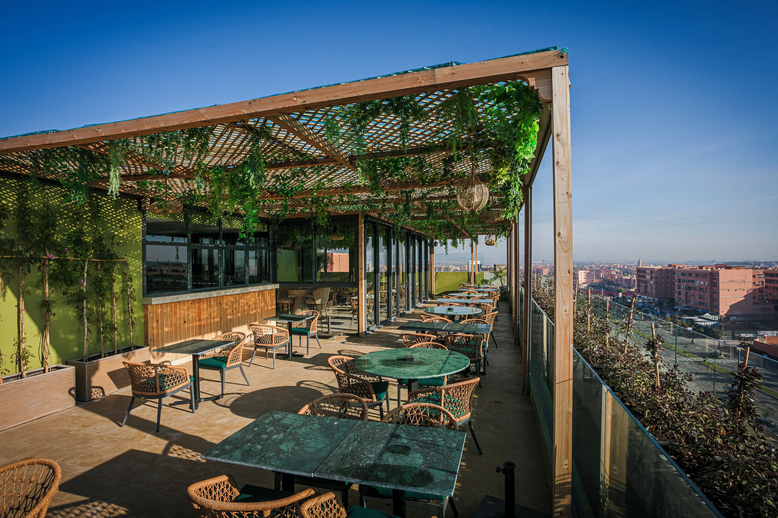 An outdoor seating area with tables and chairs, covered by a wooden pergola with hanging greenery.