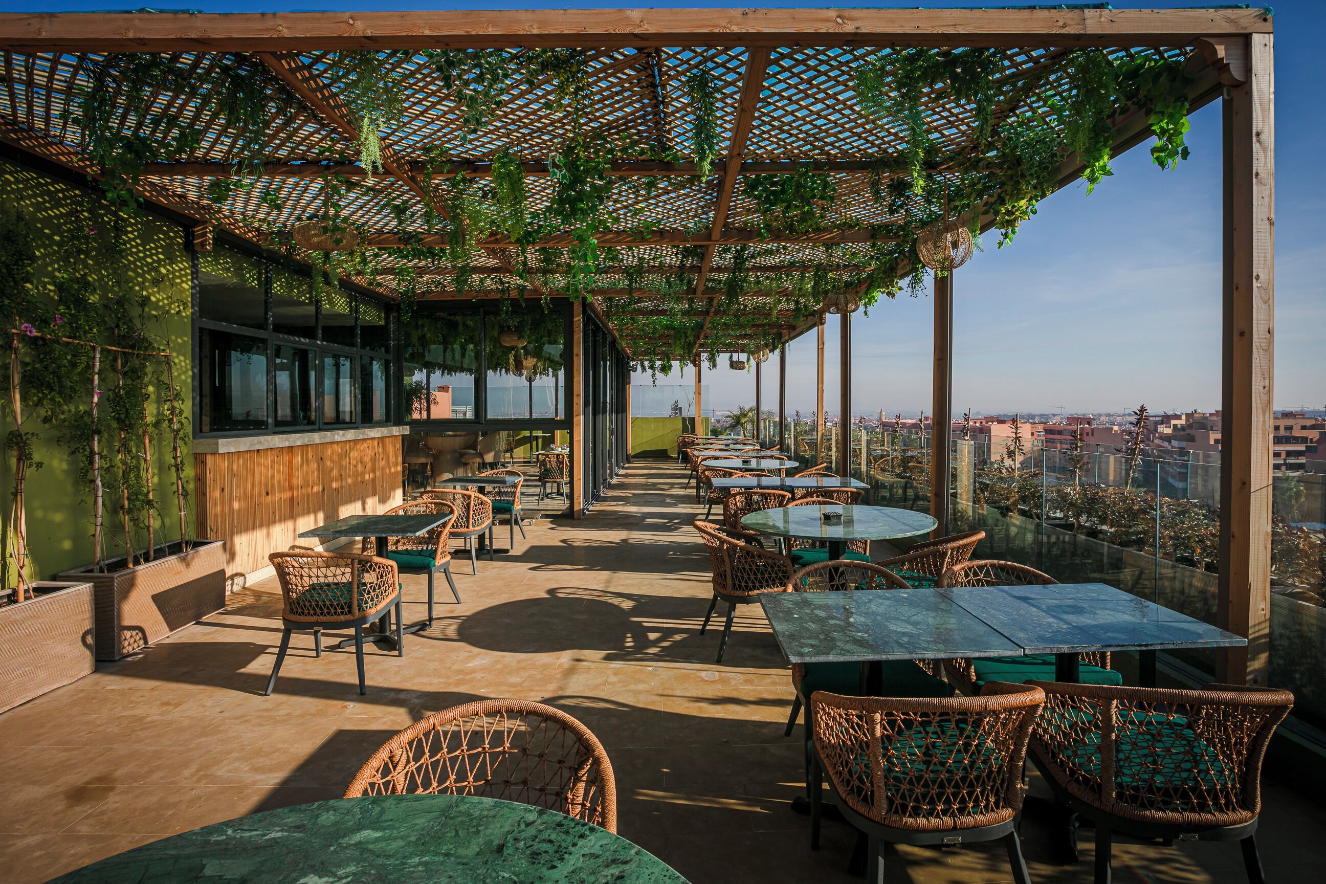 An outdoor seating area with wicker chairs and tables, covered by a wooden pergola with hanging plants, overlooking a cityscape.