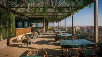 An outdoor seating area with wicker chairs and tables, covered by a wooden pergola with hanging plants, overlooking a cityscape.