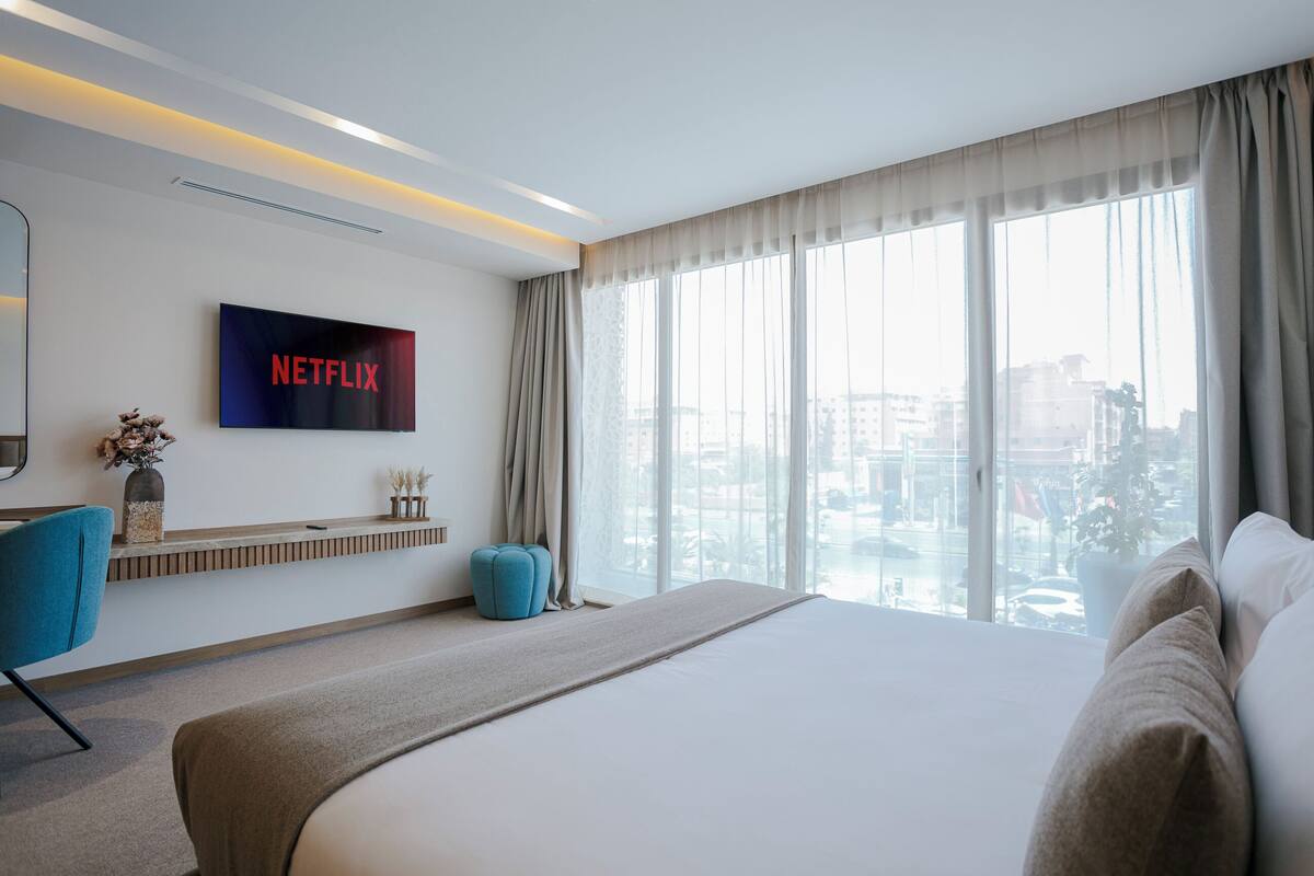 Une chambre d’hôtel moderne dotée d’un grand lit, d’un téléviseur fixé au mur, d’une vue sur la ville grâce à de grandes fenêtres, et d’un petit coin salon avec un fauteuil et un pouf.