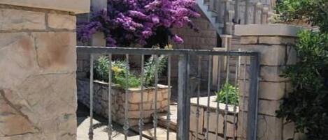 A stone building with a tiled roof, a metal gate, and purple flowers in the foreground.