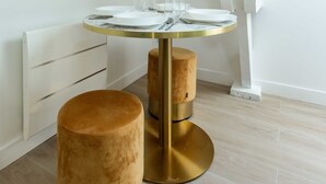 Una mesa de comedor redonda con base dorada y un reposapiés marrón, ubicada en una habitación con paredes blancas y piso de madera.