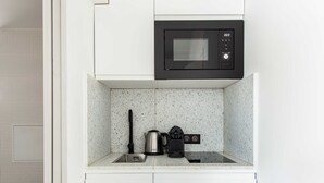 Cocina compacta con armarios blancos, un microondas negro y un hervidor de acero inoxidable sobre un salpicadero moteado.
