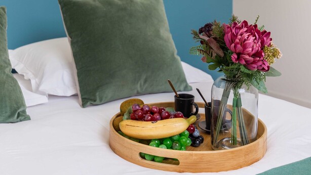 Una cama bien hecha con almohadas verdes, una bandeja con frutas y un florero, además de una pared azul con un diseño en zigzag.