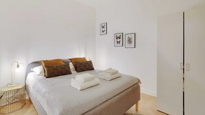 Una cama bien tendida con toallas dobladas, una lámpara de noche y un armario en una habitación minimalista con una estética limpia y blanca.