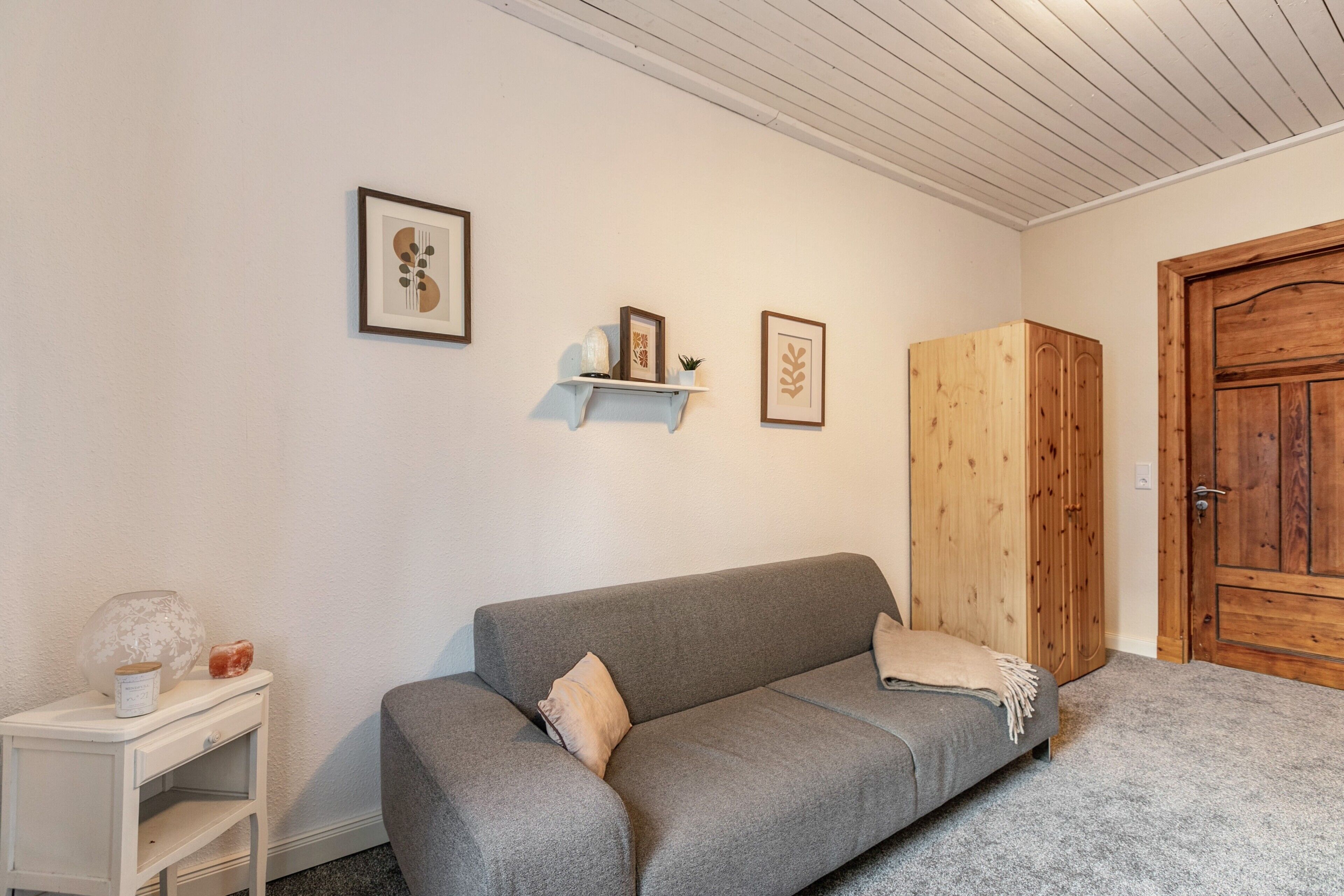 A neutral-colored room with a grey sofa, a small white nightstand, a wooden wardrobe, and two framed pictures on the wall.