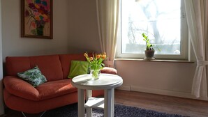 Una sala de estar con un sofá, una mesa redonda con un jarrón de flores, una ventana con cortinas y un cuadro en la pared.