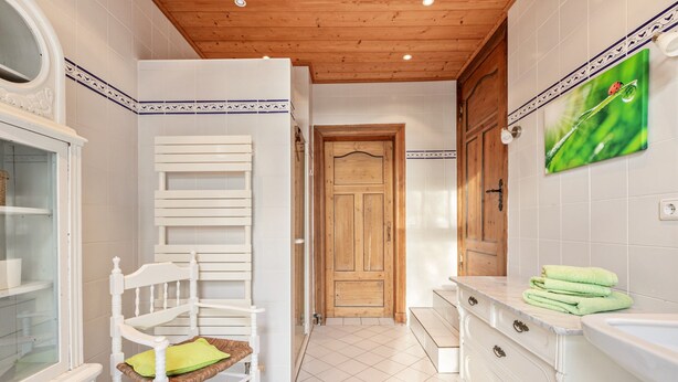 Una cocina con armarios blancos, una puerta de madera, una silla blanca con cojín verde y una planta verde en la pared.