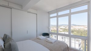 Et soveværelse med en stor seng, udsigt over bybilledet gennem store vinduer og en balkon med en stol.