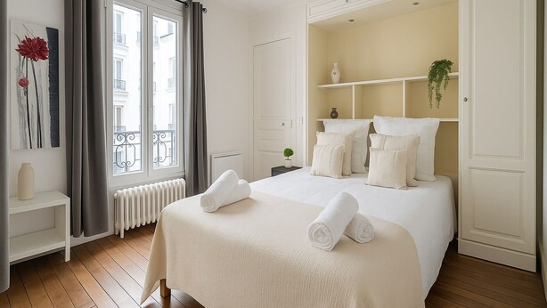 A hotel room with a large bed, a radiator, a window with curtains, a small plant, and a shelf with decorative items. - 3 pieces pres du Pantheon (Paris)